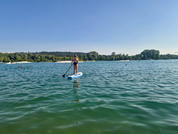 Persona en una tabla de paddle surf en un lago sosteniendo un remo; orilla arbolada y cielo azul al fondo