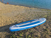 Tabla de paddle surf azul y blanca sobre una orilla arenosa; agua al fondo y algunas personas