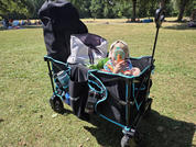 Kind met patroon zonnehoed zit in opvouwbare wagen op gras, rugzak ernaast, houdt een beker