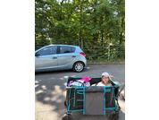 Fille assise dans une remorque pliante au bord de la route ; en arrière-plan une voiture argentée, des arbres et une clôture