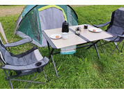Tenda verde dietro un tavolo da campeggio pieghevole con due sedie pieghevoli; sul tavolo una thermos, due piatti bianchi ciascuno con un dolcetto rotondo, un piccolo contenitore con cucchiaio e un panno sotto il tavolo; erba verde