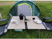 Tavolo pieghevole davanti a una tenda verde sull'erba con due piatti ciascuno con un biscotto rotondo, due tazze, una caffettiera, una French press, un vasetto di burro e due sedie da campeggio