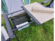 Vista parziale di un tavolo con un pannello in legno chiaro accanto a un telaio metallico nero; sotto una tasca in tessuto nero con un panno bianco e un cuscino colorato; erba verde, parete di una tenda sullo sfondo e bracciolo di una sedia pieghevole a destra