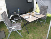 Zwei gepolsterte Campingstühle und ein klappbarer Tisch unter dem Wohnwagenvorzelt; auf dem Tisch zwei Teller auf Platzsets, eine Laterne, eine Tasse und eine Wasserflasche; Grasboden