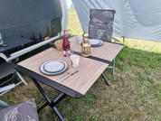 Klappcampingtisch mit Holzoptik, zwei Teller, eine Tasse, Besteck, rote Flasche, Laterne, zwei gepolsterte Klappstühle, Gras und Teil eines Wohnwagenvorzelts