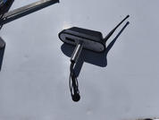 Dispositivo negro con dos barras delgadas, conectado a un enchufe cilíndrico plateado con cable, sobre una superficie gris claro brillante; sombra visible