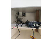 Aparato negro con cuatro puertos Ethernet en una estantería, varios cables negros detrás, caja de enchufe blanca en la pared posterior, destornillador en la superficie inferior, tubo marrón a la derecha