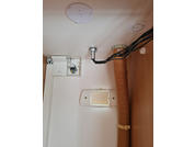 Intérieur avec plafond blanc et plaque de fixation ronde, petit boîtier blanc ouvert avec fils visibles, faisceau de câbles noirs, tuyau isolé brun à droite et ouverture murale rectangulaire