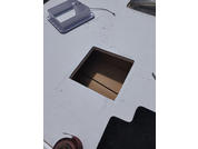 Superficie piatta bianca con un'apertura quadrata che mostra cartone marrone; in alto a sinistra un elemento in plastica bianca con un cavo allentato, in alto a destra un pezzo metallico rettangolare, in basso a destra tappeto nero, in basso a sinistra una cinghia marrone e piccole particelle di polvere intorno all'apertura