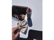 Mano che tiene un panno beige chiaro accartocciato su una superficie bianca, oggetto rettangolare nero lucido che riflette le dita, bomboletta spray con tappo rosso, tappetino nero strutturato in basso a destra