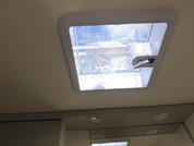 Finestra rettangolare nel soffitto con cornice bianca e maniglia laterale, con vista su cielo nuvoloso; sotto, soffitto con tre faretti rotondi incassati e uno specchio che riflette la finestra