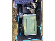 Blick von oben in einen offenen schwarzen Wagen mit türkisfarbener Einfassung auf Gras, mit grüner Kühlbox, grüner Trinkflasche, blau-grauer Sandale, gefalteter Kleidung und einer schwarzen Tasche