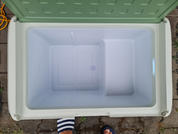 Glacière ouverte et vide avec couvercle vert clair et intérieur blanc ; en haut à gauche un tuyau jaune, en bas des pieds partiellement visibles portant des sandales rayées bleu et blanc sur pavé