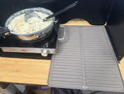 Tragbare Elektrokochplatte auf Holzfläche, darauf runde Aluminiumform mit hell-cremiger Masse und schwarzem Löffel; rechts graue gerillte Silikonmatte; schwarzer Windschutz im Hintergrund