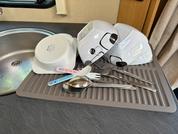 Grau gerillte Abtropfmatte auf Küchenarbeitsplatte neben Spüle, darauf zwei weiße Kunststoffschalen, zwei bunte Plastiklöffel (rosa, blau), eine kleine Edelstahlschüssel, ein Metalllöffel und zwei Metallgabeln