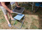 Lower body of a person placing a square tray on a blue folding stool on dry grass; camping chairs and tent poles in the background