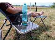 Part of a person sits in a folding camping chair with legs stretched on a footrest; a large plastic water bottle and a glass are on a small folding table in front; grass field and distant landscape in the background