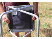 Two hands hold a folded camping chair with a gray metal frame and black fabric seat, grass in the background