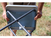 Hands holding a folded chair with a gray metal frame and blue fabric seat, grass in the background