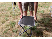 Lower body of a standing person wearing shorts; hands hold a small folding stool with metal frame and dark square seat on grass