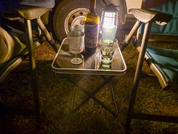 Folding table between two camping chairs with a wine bottle, two wine glasses, a tumbler and a lantern; vehicle wheel visible in background on grass