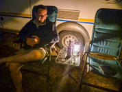 A seated man wearing glasses in a camping chair next to a caravan wheel; a small table with a lantern, a water bottle and a thermos; an empty camping chair beside him