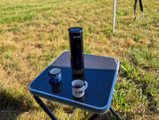 Square folding table with black top on grass. On the table a black thermos and two cups (one dark blue, one white with blue pattern). Tripod legs visible in the background