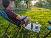 Person sitting in a folding lawn chair on grass; small table with a wine bottle and a can; fenced field and a brown animal in the distance