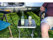 Small black folding table with a glass bottle and a can, two camping chairs, person on the right wearing a burgundy shirt and light-colored shorts, open trunk of a white vehicle with shoes inside, green grass