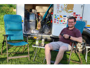 Man with glasses sitting in a camping chair in front of an open camper; small table with two cans and a water bottle; empty camping chair beside him
