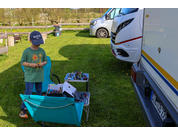 A child wearing a baseball cap and green T‑shirt stands on grass next to two teal folding chairs and two small tables with several small models; a white camper van is on the right