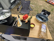 Wooden table with gas stove and lidded cooking pot, frying pan with two green zucchinis, metal spatula and wooden turner, knife, red bell pepper, onion, cucumber, small dish with butter, bag of pasta, two camping chairs and tent in the background