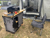 Portable camping kitchen unit with wooden countertop, black frying pan and pot, bread roll and red bell pepper on the counter; beside it a gray folding camp chair; a gray tent in the background