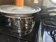Stainless steel pot with gray lid and light wooden knob on a gas stove; a black pan to the right