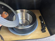 Wooden shelf with nested stainless steel bowl, glass lid with brown knob (held by a hand on the left), blue silicone mat underneath, black-handled tool on the right, dark interior walls