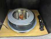 Round stainless-steel pot with lid on a blue bowl, both on a light wooden surface; to the right a long black device with a white button