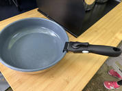 Gray frying pan with black handle on a wooden surface; metal rack in the background and pink shoes at the right edge