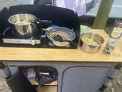 On a wooden countertop: an electric hotplate with a pot, a frying pan with a spatula, a stainless steel bowl with pasta, and a beverage can; in the background a cloth, a tree trunk, and a parked van
