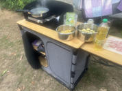 Foldable camping kitchen cabinet with wooden countertop; frying pan on gas burner with steam; two metal bowls with round light-colored slices; yellow plastic bottle with liquid; plate with pink slices; purple bag in background; grassy ground