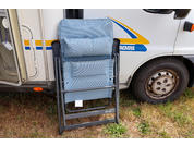 Sedia da campeggio piegata azzurro chiaro appoggiata al fianco di un camper bianco con strisce blu e gialle, accanto a una ruota su erba secca