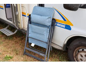 Sedia pieghevole imbottita azzurro chiaro, piegata, con poggiatesta e struttura nera appoggiata al fianco di un camper bianco con striscia blu-gialla e scritta parziale visibile; porta aperta, gradino e prato visibili