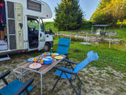 Camper con porta aperta su prato accanto a uno stagno; tavolo pieghevole con piatti, tazze, una ciotola e posate e sedie pieghevoli blu; pontile di legno e tettoia sullo stagno; alberi sullo sfondo