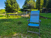 Poltrona pieghevole da giardino blu sul prato davanti a uno stagno con passerella in legno e un riparo coperto, alberi e cielo azzurro sullo sfondo