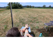 Rückenansicht einer sitzenden Person mit ausgestreckten Beinen und Füßen auf einem hellblauen Stoff, dahinter ein trockenes Grasfeld mit vereinzelten Bäumen, rechts ein klappbarer Tisch und eine grüne Plastikflasche, blauer Himmel