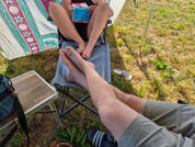 Zwei Personen, nur Beine und nackte Füße sichtbar; eine hält ein blau-rotes rechteckiges Gerät; gemusterter Stoff als Sonnenschutz, Gras und Sandalen am Boden
