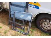 Zwei zusammengeklappte hellblaue Klappstühle lehnen an der Seite eines weißen Wohnmobils neben dessen hinterem Rad auf trockenem Gras