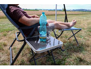 Person in Campingstuhl mit hochgelegten Beinen, Füße sichtbar; auf kleinem Tisch eine blaue Plastikflasche und ein Glas; Wiese im Hintergrund