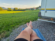Blick auf nackte Unterschenkel und Füße in schwarzen Shorts auf blauem Liegetuch; rechts ein weißer Wohnwagen mit blauen und gelben Streifen und ein kleiner Klapptisch; Kies, grüne Wiese, Dorf und Berge unter blauem Himmel