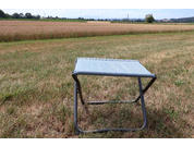 Faltbarer Hocker mit hellblauem Stoffsitz und dunklem Metallgestell auf trockenem Gras; im Hintergrund Getreidefeld, vereinzelte Bäume und Horizont