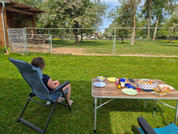 Person sitzt in klappbarem Gartenstuhl auf Rasen, Rücken zur Kamera; rechts Campingtisch mit Tellern, Würzflaschen und Brot; im Hintergrund Bauzaun, Bäume und Holzschuppen